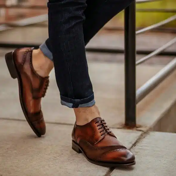 A close-up of a person's legs and feet wearing dark blue jeans with the cuffs rolled up and brown leather wingtip dress shoes.