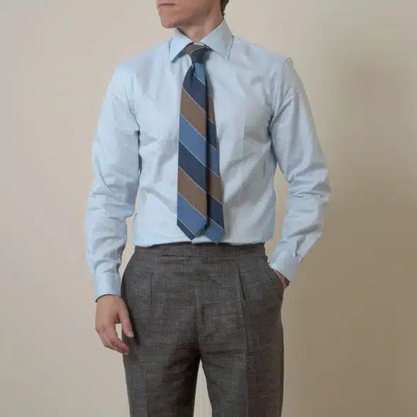 Man dressed in a light blue dress shirt with a wide striped blue and brown tie, paired with grey dress pants, standing against a plain background with one hand in his pocket.