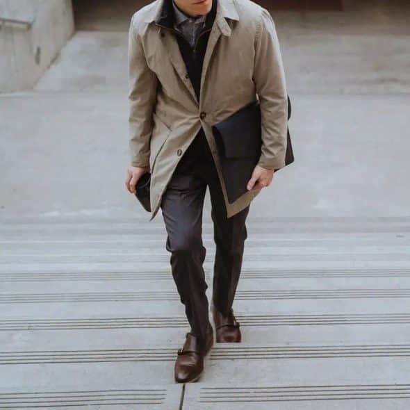 A man wearing a light brown coat over dark layers, with dark trousers and brown double-monk strap shoes, carrying a black bag and a cup, walking up concrete stairs.