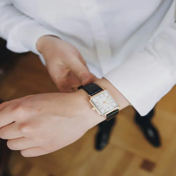 A person in a white shirt adjusting a gold rectangular wristwatch with a black strap.
